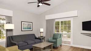 A living room with a grey sofa, a green armchair, a wooden coffee table, a ceiling fan, a TV mounted on the wall, and a large window with curtains. - Club Wyndham at the Cottages (North Myrtle Beach)