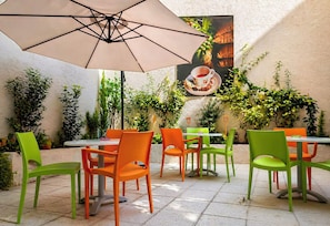 Eine Außenpatio mit Tischen und Stühlen, umgeben von viel Grün und einem an der Wand hängenden Bild.