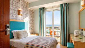 A hotel room with a bed, a desk, a chair, a balcony with a view, and a wall with a pattern. - City Life Port Hotel (Heraklion)