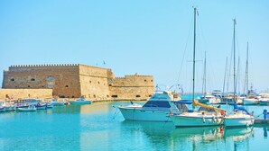 A marina with boats docked near a historic stone fortress. - City Life Port Hotel (Heraklion)