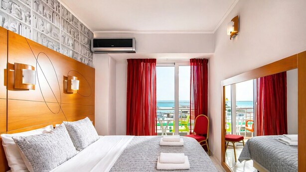 A hotel room with a bed, a TV, a balcony with red curtains, and a view of the sea. - City Life Port Hotel (Heraklion)