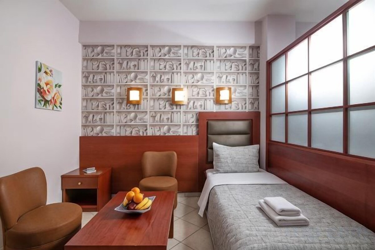 A hotel room with a bed, a desk with fruit, two chairs, a nightstand, and a wall with a patterned design.