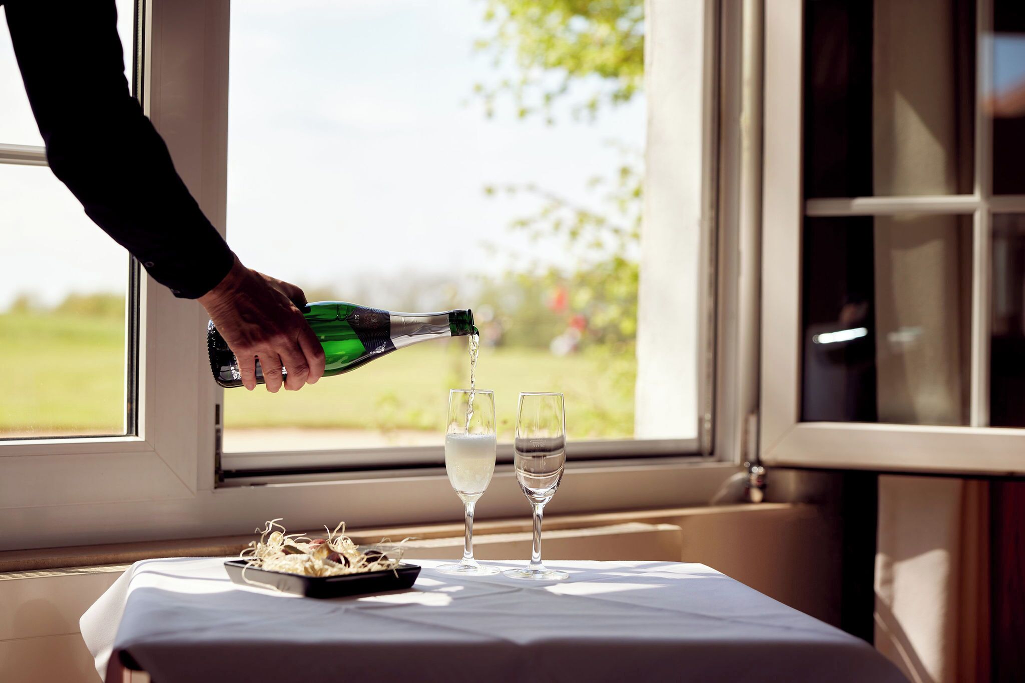 A bottle being poured into two glasses on a table by a window.