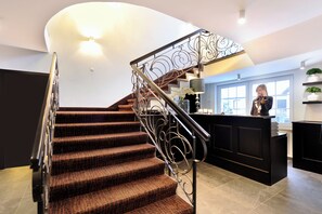 Un escalier recouvert d’une moquette à motifs et doté d’une rampe en métal, menant à un comptoir d’accueil agrémenté d’une plante et d’une fenêtre.