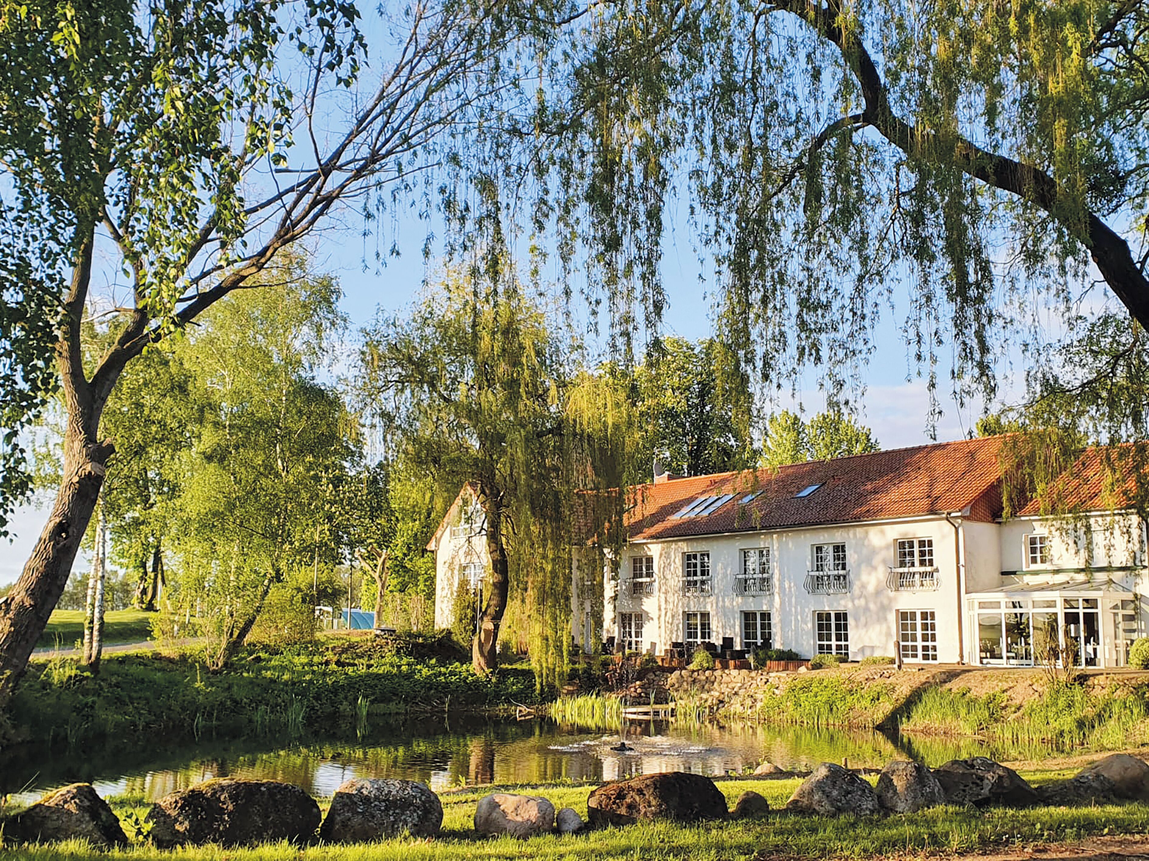 A large white building with multiple windows and a red roof, surrounded by trees and a pond.