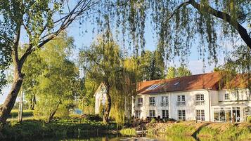 A large white building with multiple windows and a red roof, surrounded by trees and a pond.