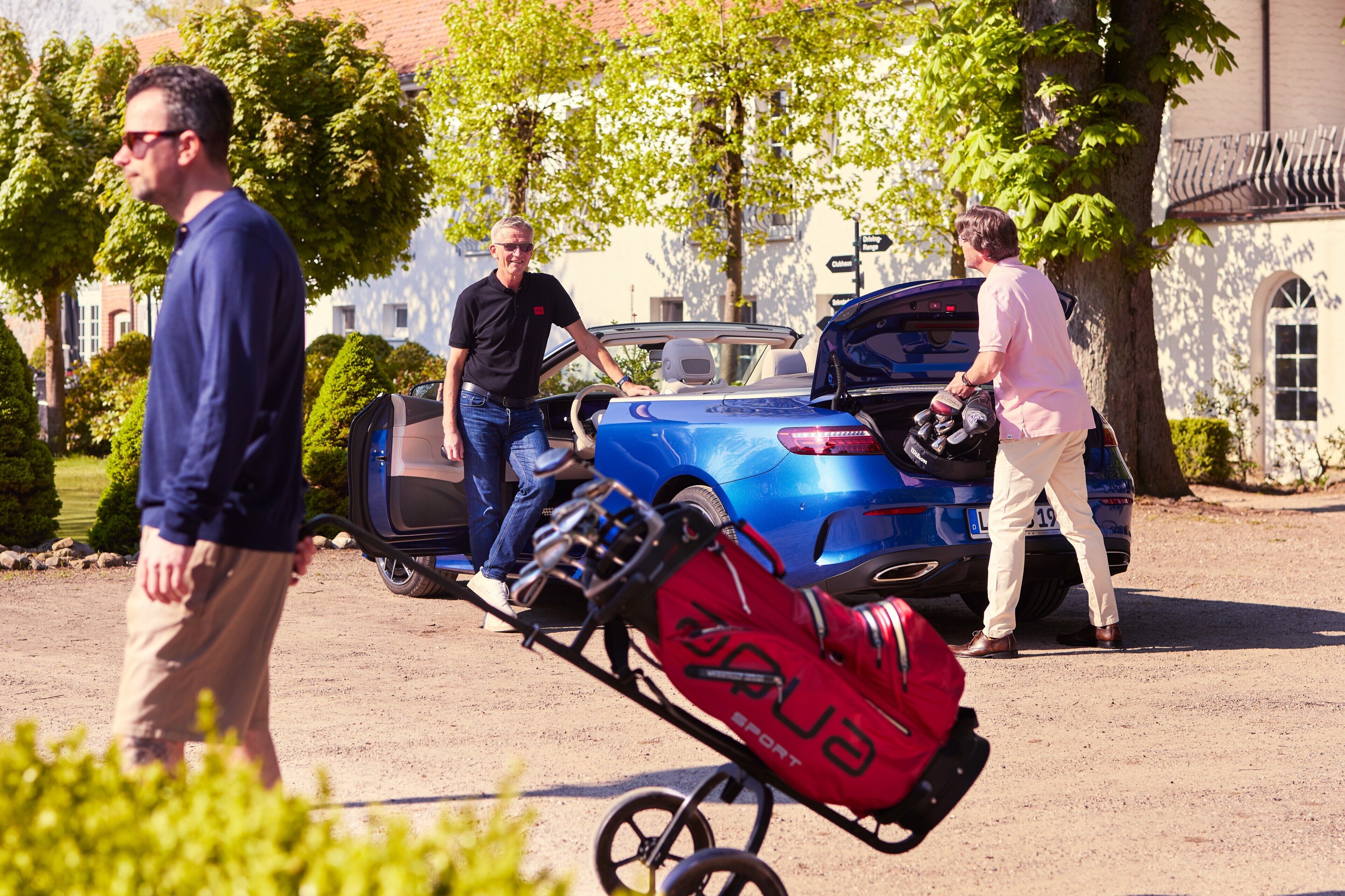 A blue convertible with its trunk open, a red bag labeled 'SPORT' on a cart, and a person in a pink shirt standing by the vehicle.