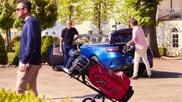 A blue convertible with its trunk open, a red bag labeled 'SPORT' on a cart, and a person in a pink shirt standing by the vehicle.