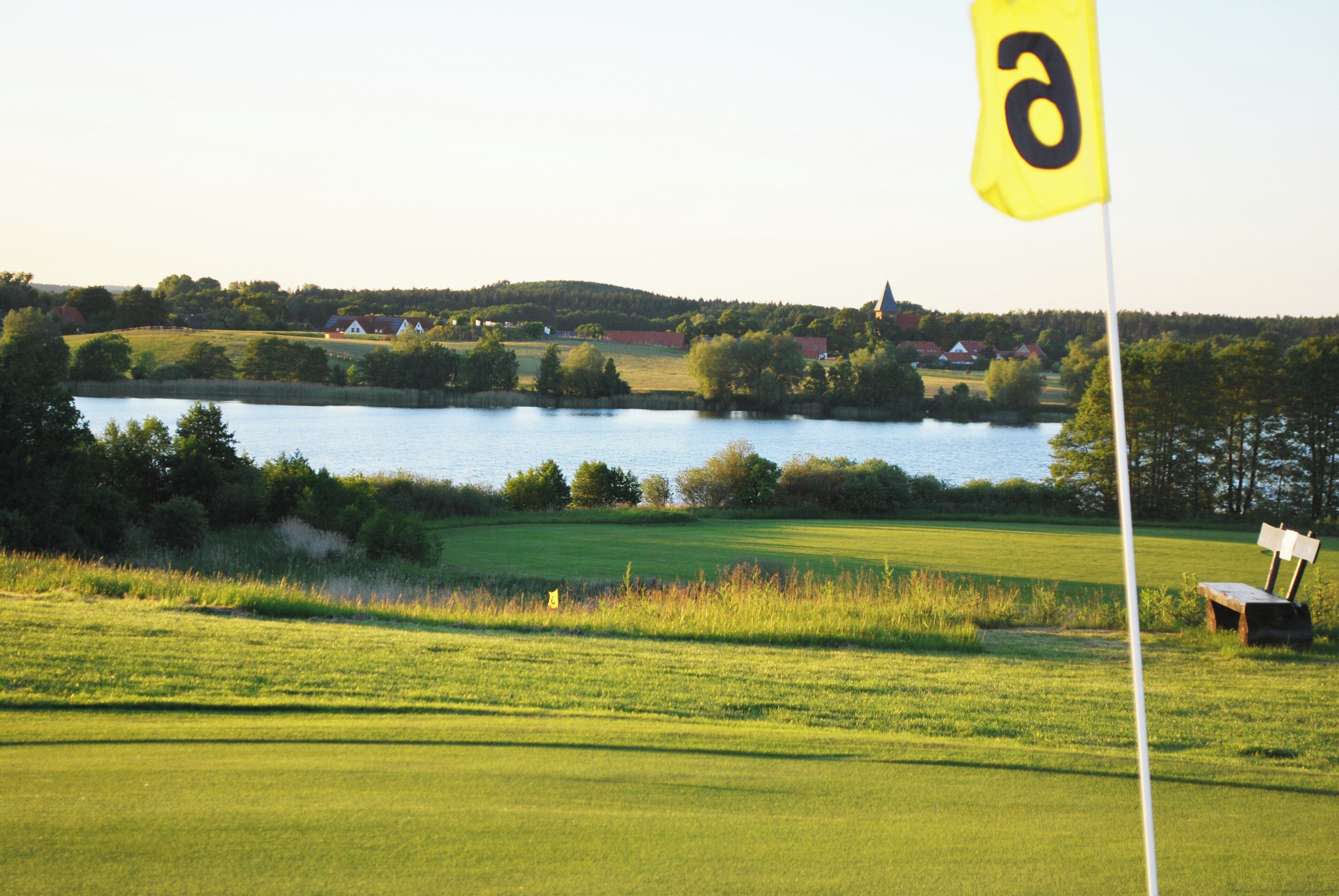 A golf course with a flag, a lake, and houses in the background.