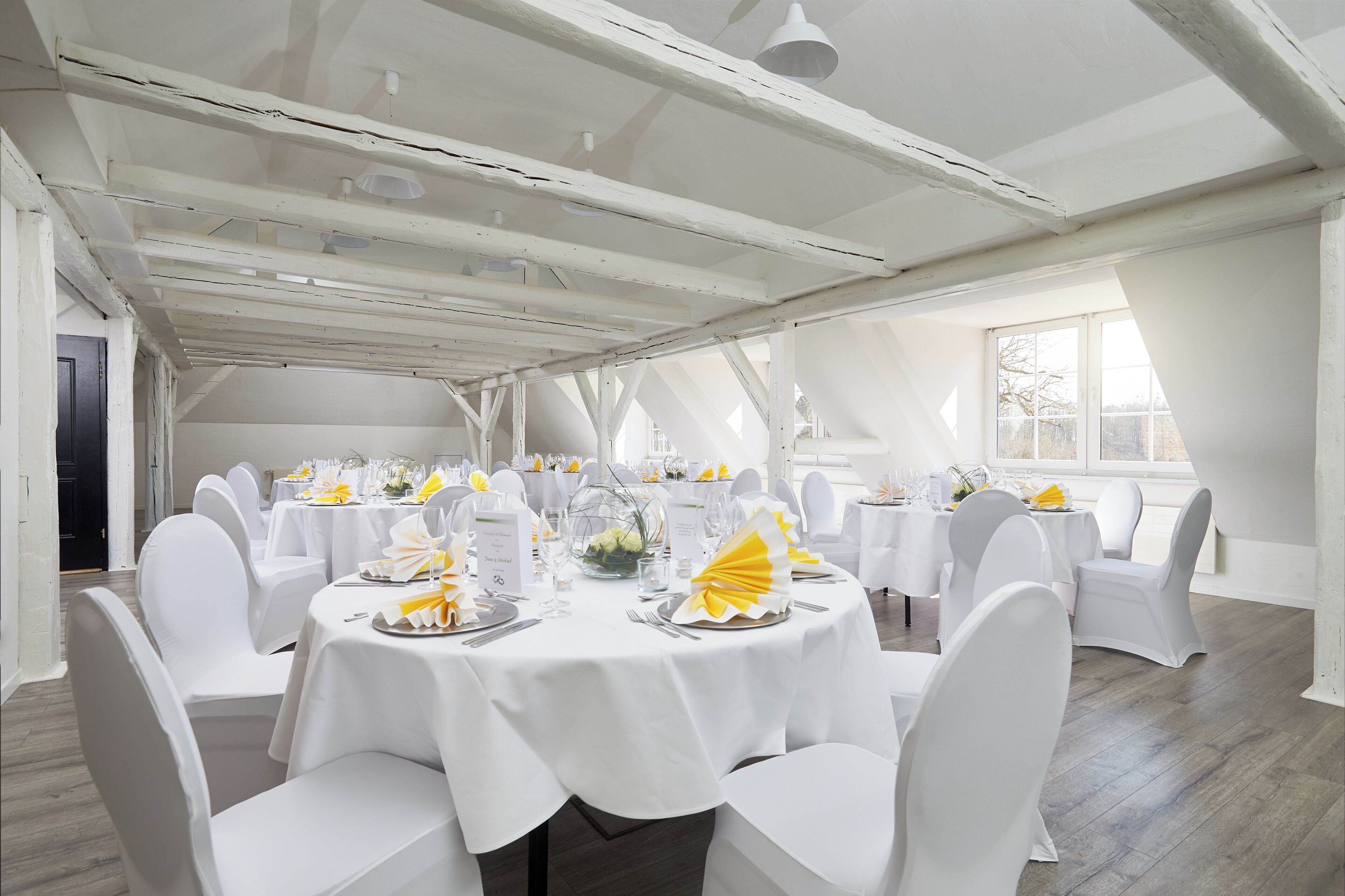 A large room with multiple round tables, white chairs, and a high ceiling with exposed beams.