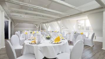 A large room with multiple round tables, white chairs, and a high ceiling with exposed beams.