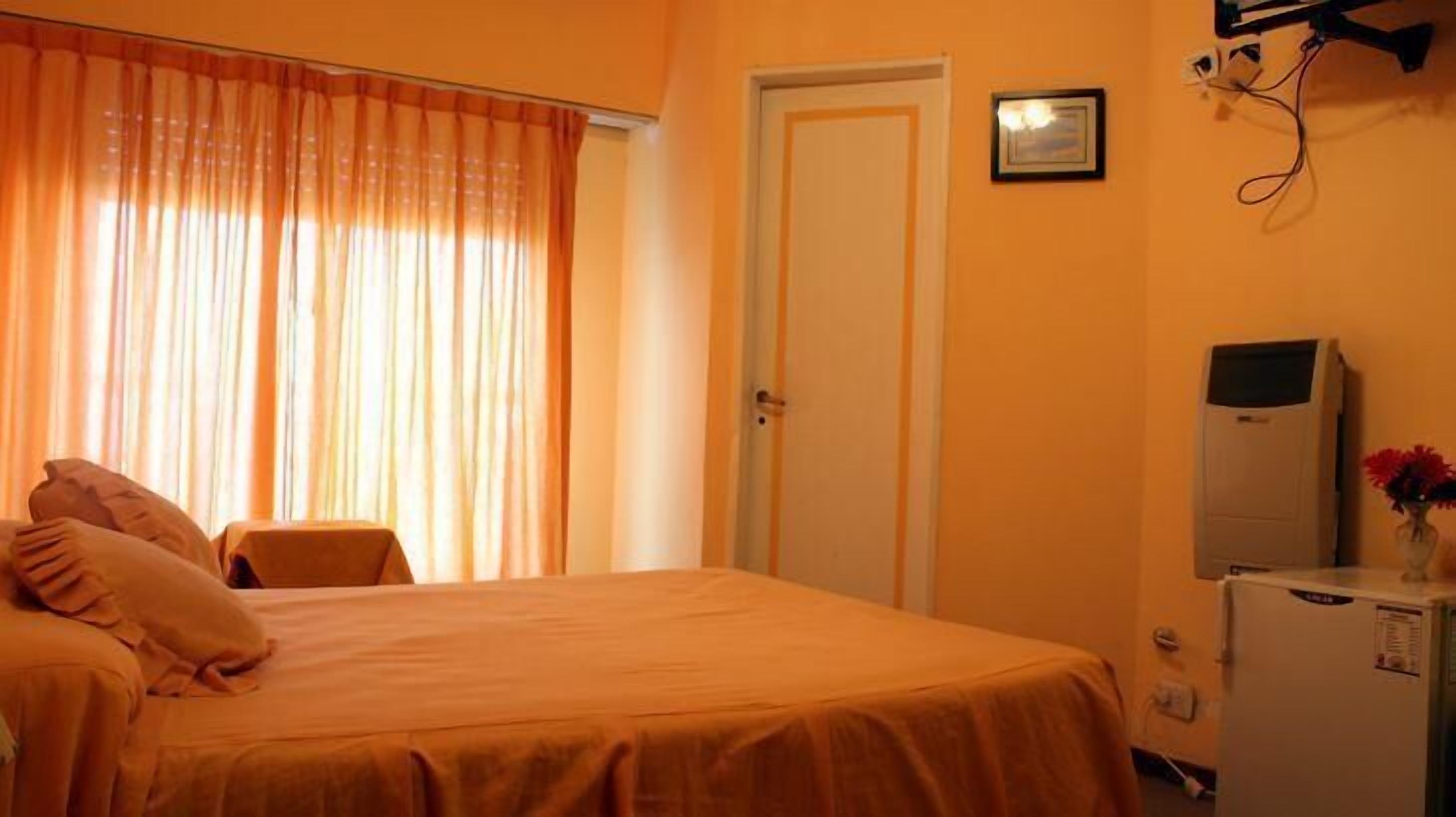A room with a bed, a door, a small table with a vase, and a refrigerator.