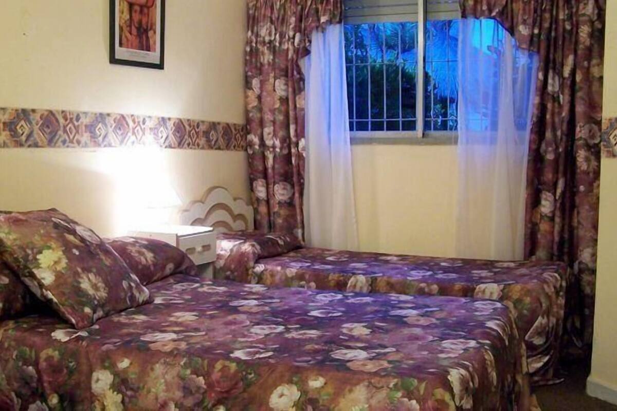 Een slaapkamer met twee bedden, een nachtkastje, een lamp en een raam met gordijnen.