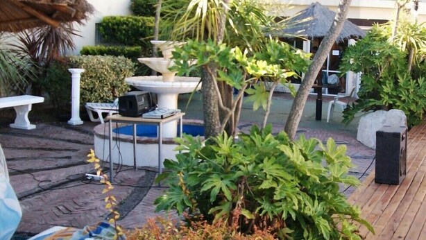 An outdoor patio area with a white table and chairs, a stone fountain, and various plants. - San Remo Resort Hotel (Santa Teresita)