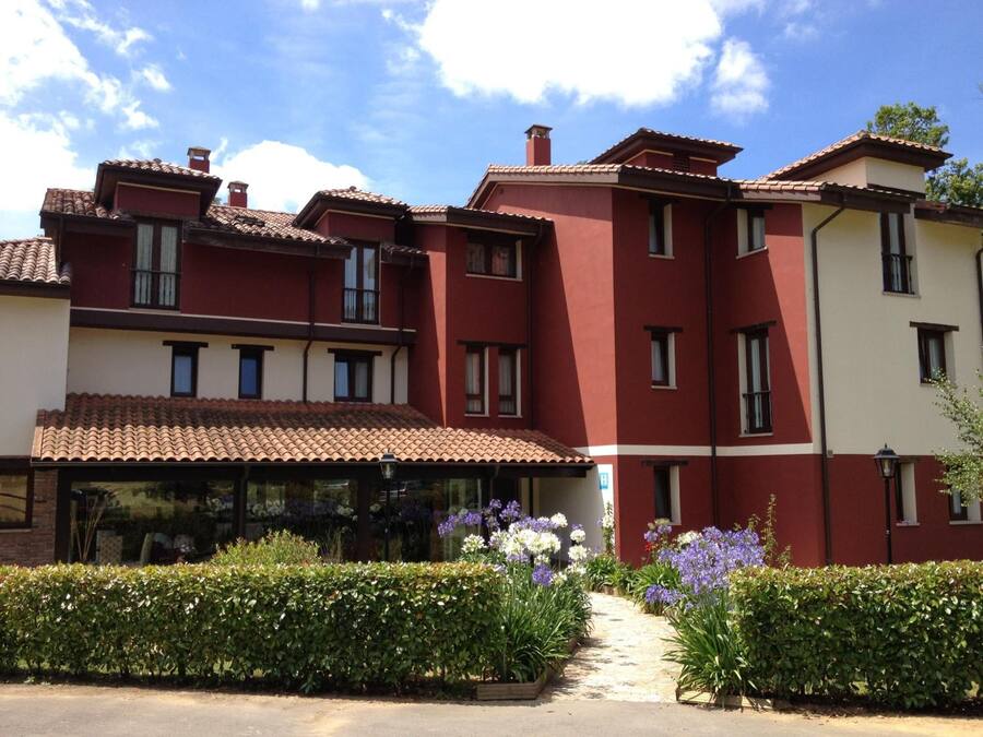 Un edificio de varios pisos con fachada roja y blanca, techo de tejas y un jardín con diversas plantas y flores.