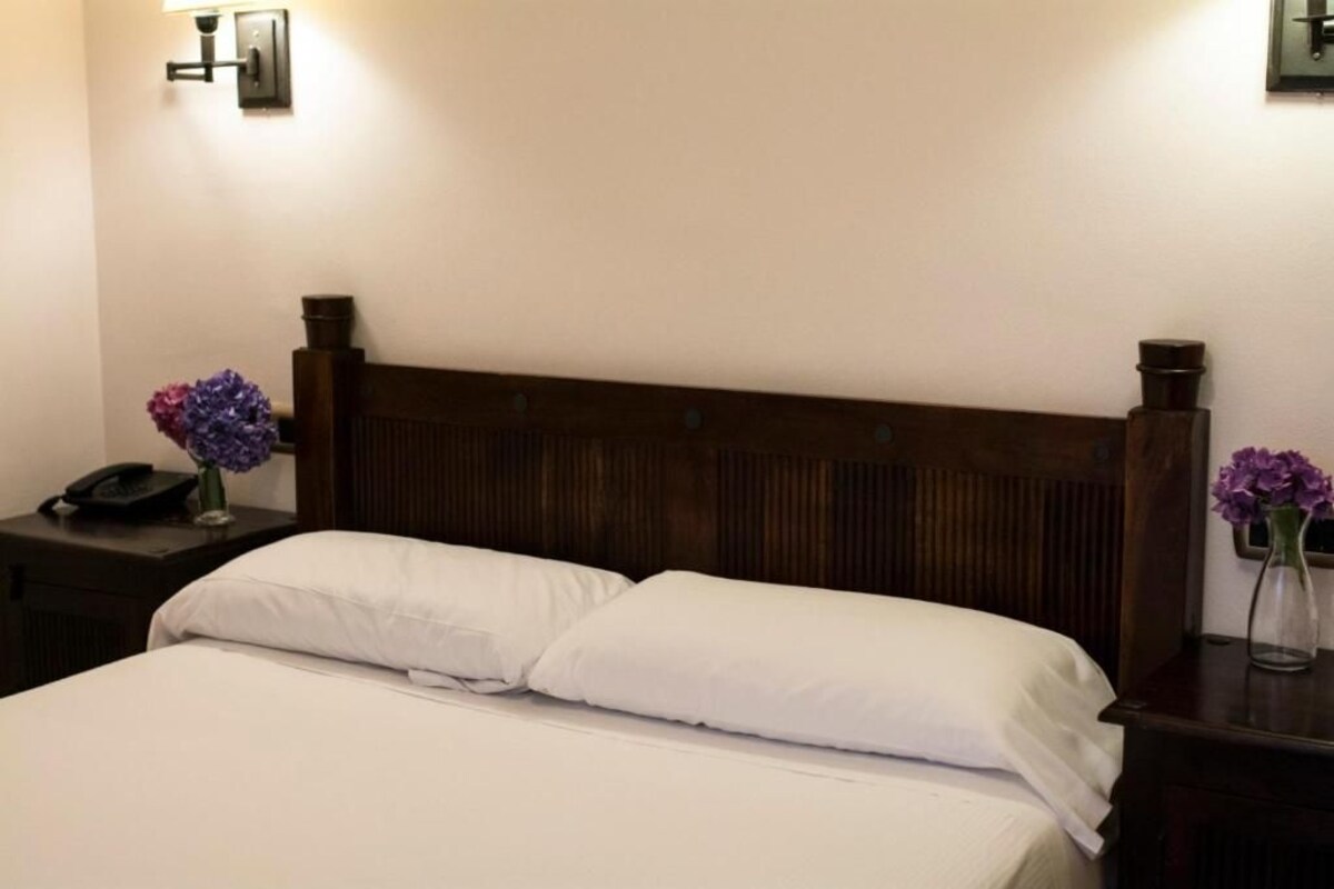 Una cama bien tendida con sábanas blancas, cabecera de madera y dos mesitas de noche con flores moradas.
