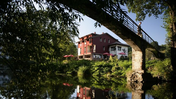 Un edificio de dos plantas con fachada roja, un puente de piedra y un reflejo en el agua.