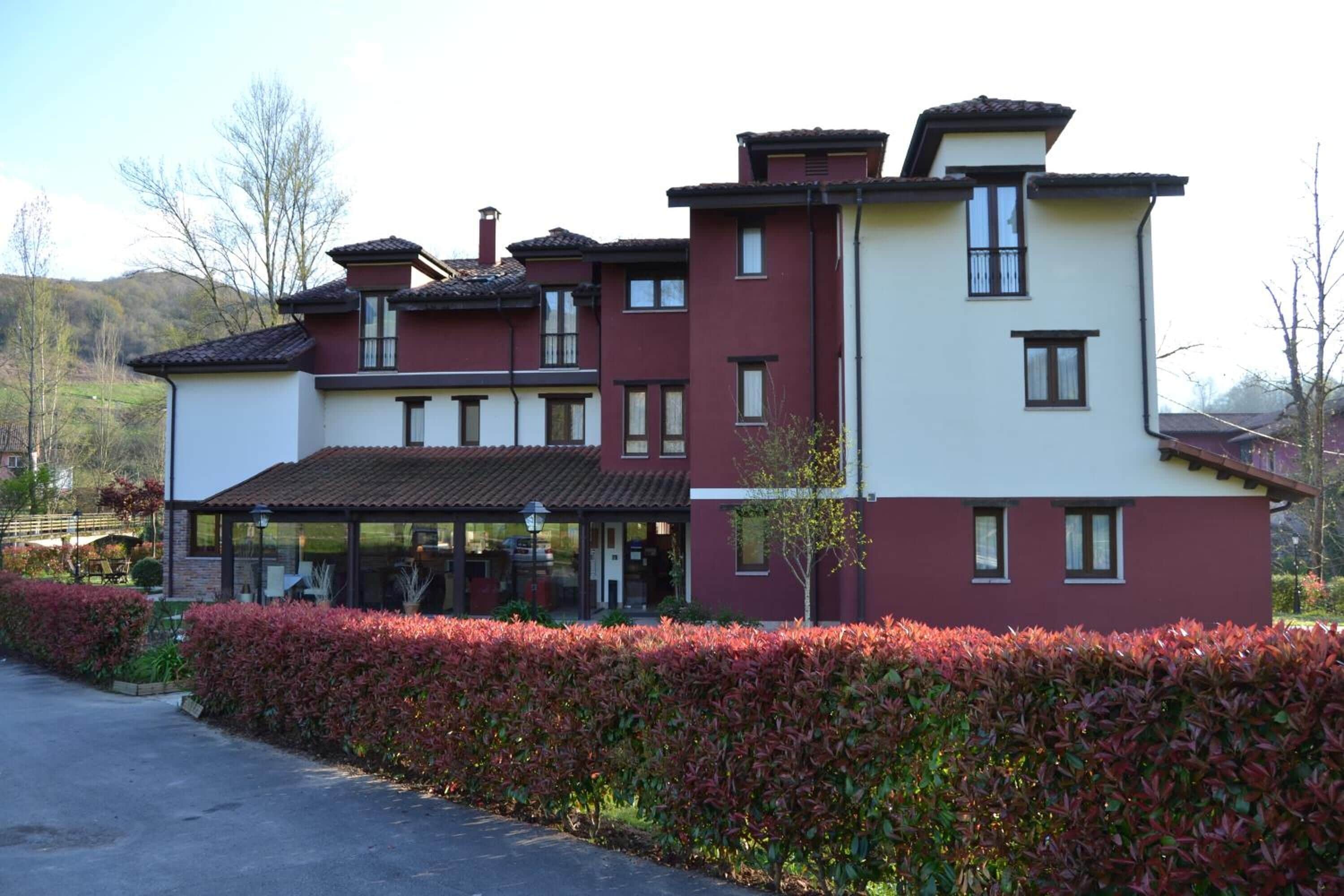 Un edificio de varios pisos con paredes exteriores en tonos rojo y blanco, un área de entrada cubierta y un seto bien cuidado al frente.