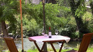 Una mesa redonda de madera con mantel blanco, dos sillas de madera, una botella de vino y dos copas de vino colocadas junto a un estanque.