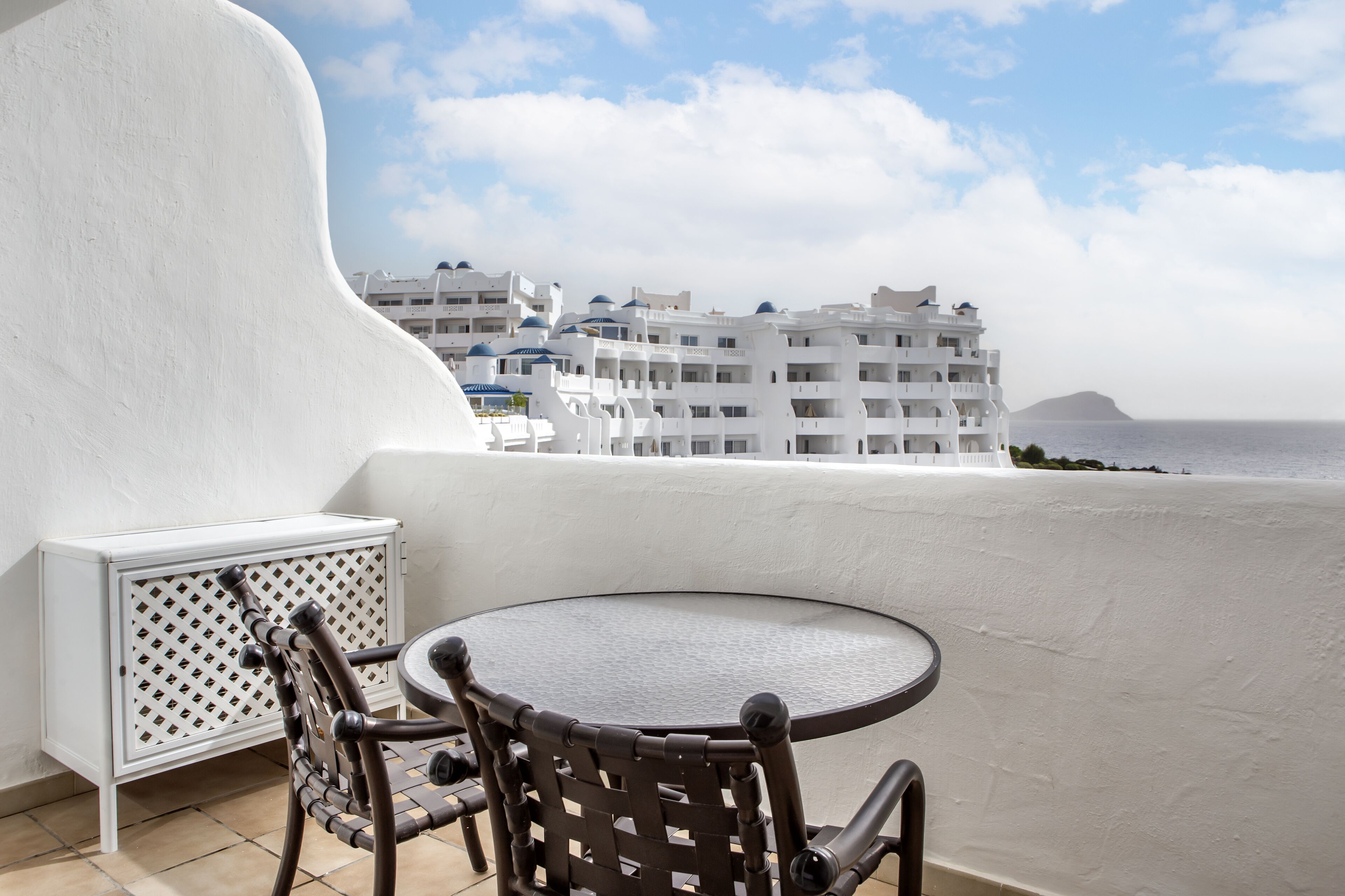 A balcony with a round table and two chairs, overlooking a coastal area with white buildings and a clear sky.