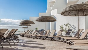 Une rangée de chaises longues, surmontées de parasols en osier, est disposée sur une terrasse carrelée, avec un bâtiment blanc en arrière-plan.