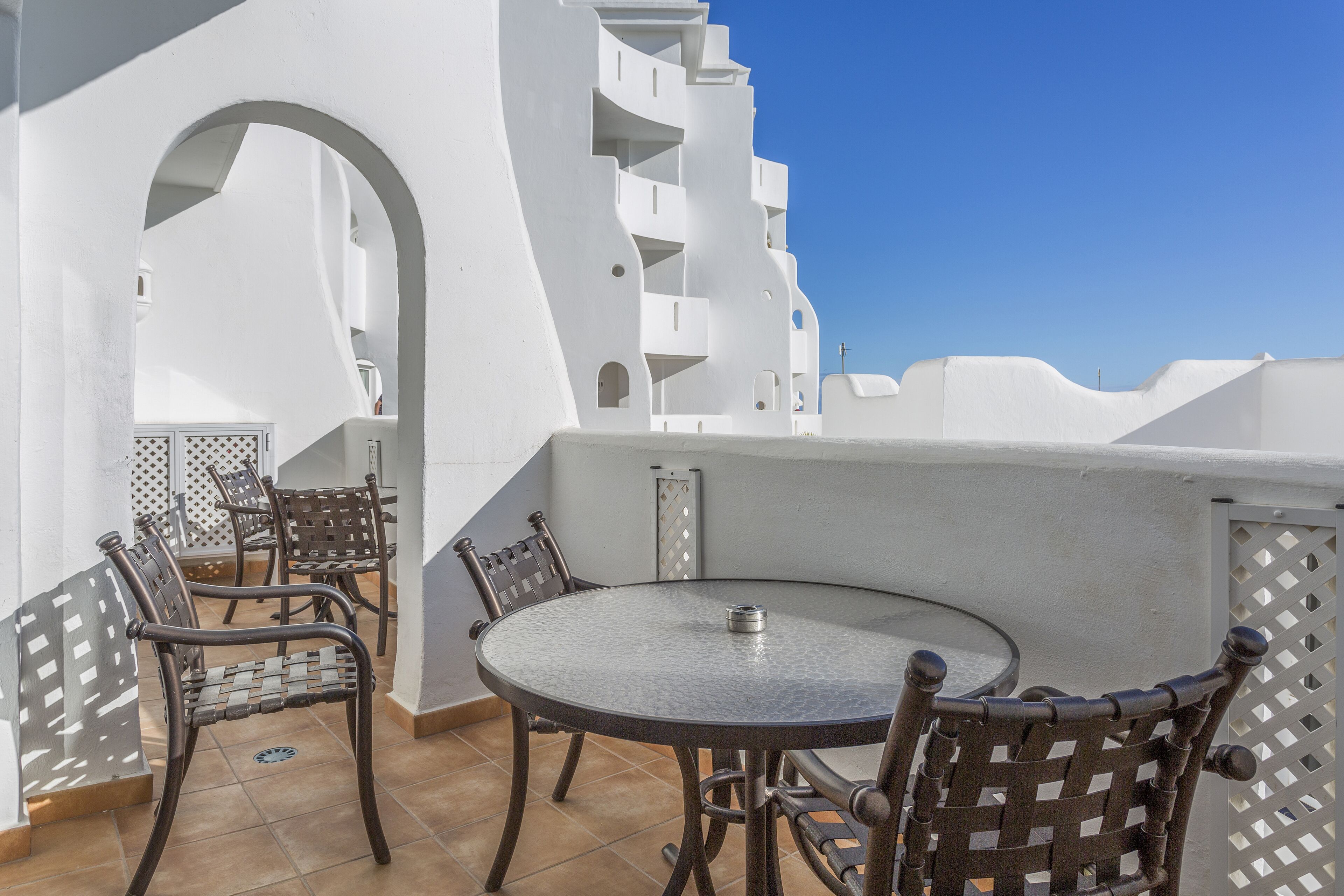Une terrasse avec une table ronde et quatre chaises, entourée de murs blancs et surplombant un ciel d’un bleu limpide.