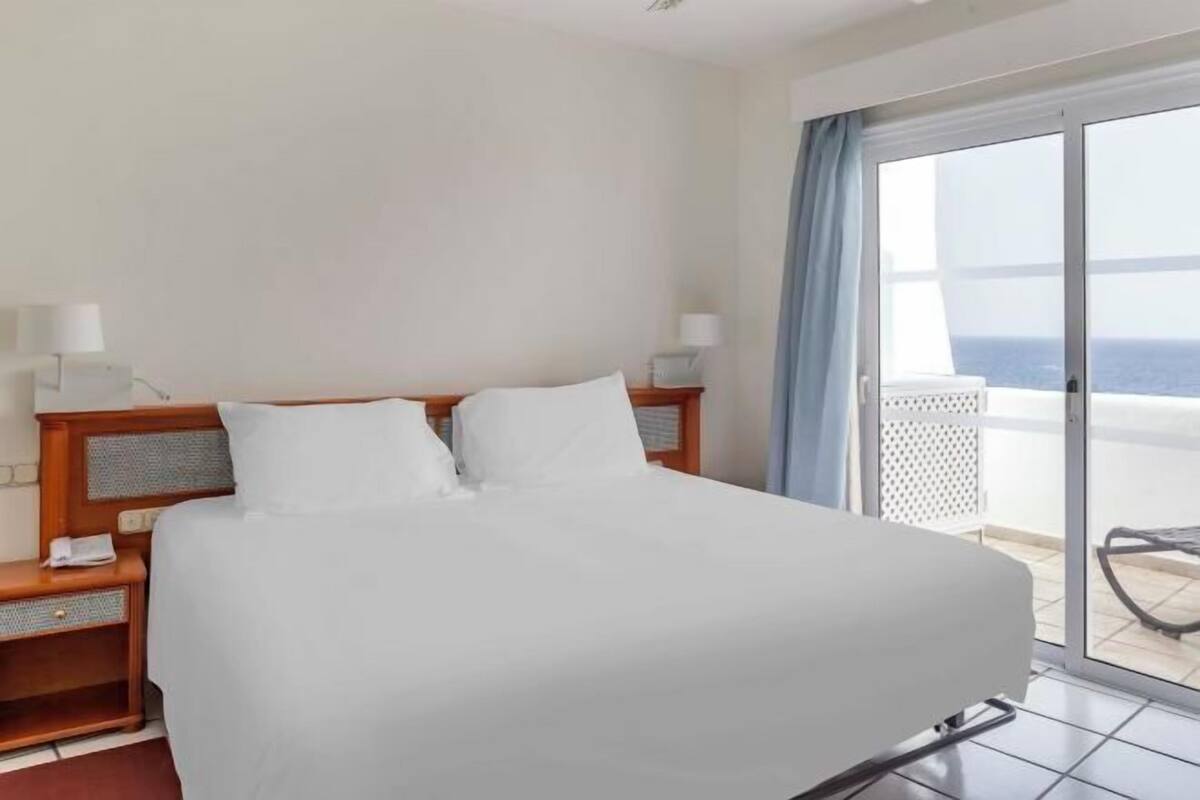 Une chambre d’hôtel avec un lit, une table de chevet, une lampe et une vue sur l’océan, visible à travers une porte-fenêtre coulissante.