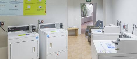 Un cuarto de lavandería con varias lavadoras y secadoras, un cartel con instrucciones de uso y una silla de mimbre al fondo.