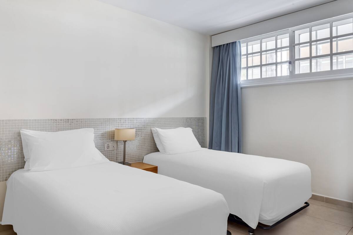 Une chambre d’hôtel avec deux lits, une table de chevet, une lampe et une fenêtre avec des rideaux.