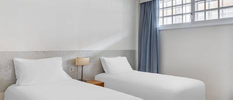 A hotel room with two beds, a nightstand, a lamp, and a window with curtains.