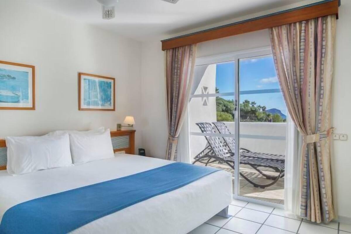 Une chambre d’hôtel avec un lit, une table de chevet, une lampe, un balcon avec une chaise et une vue sur la mer.