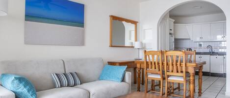 Un salon comprenant un canapé, une table basse et un coin repas avec des chaises et une table. On peut apercevoir la cuisine à travers une arche.