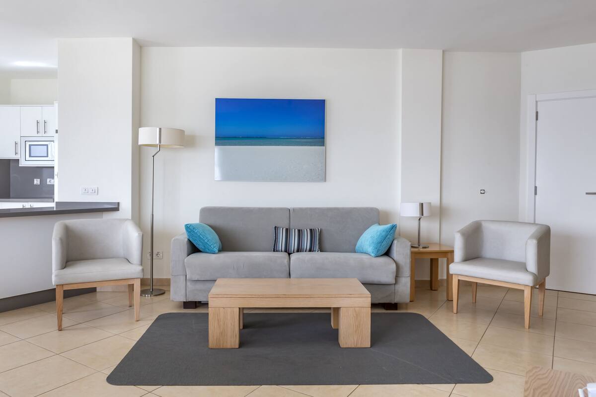 Un salon moderne avec un canapé gris, deux fauteuils, une table basse en bois et un tableau représentant une scène de plage.