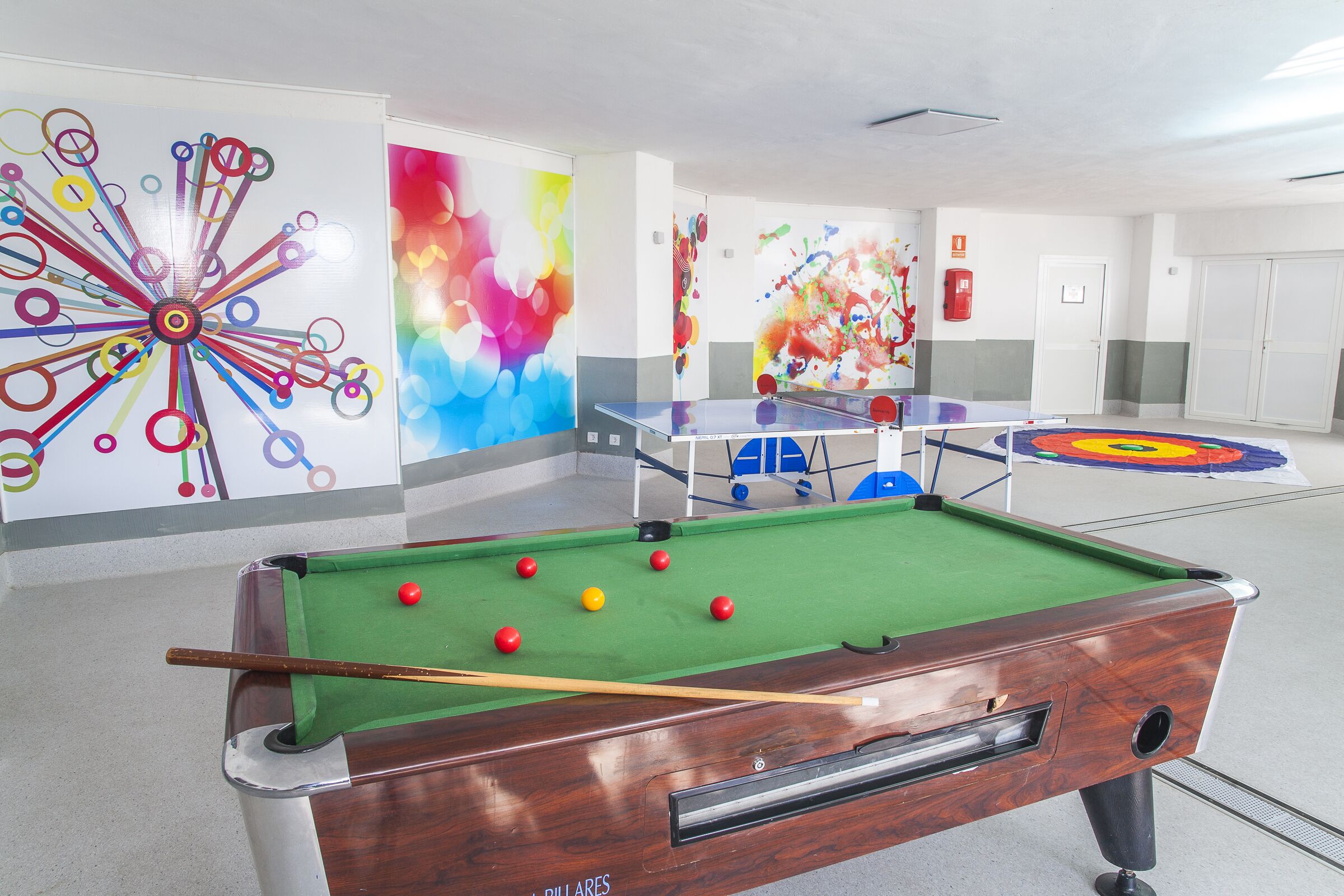 Una mesa de billar con bolas, un dardo y arte mural colorido en una habitación con paredes blancas y una puerta al fondo.