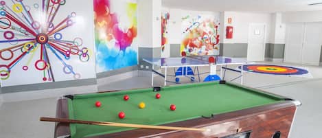 A pool table with balls, a dartboard, and colorful wall art in a room with white walls and a door in the background.