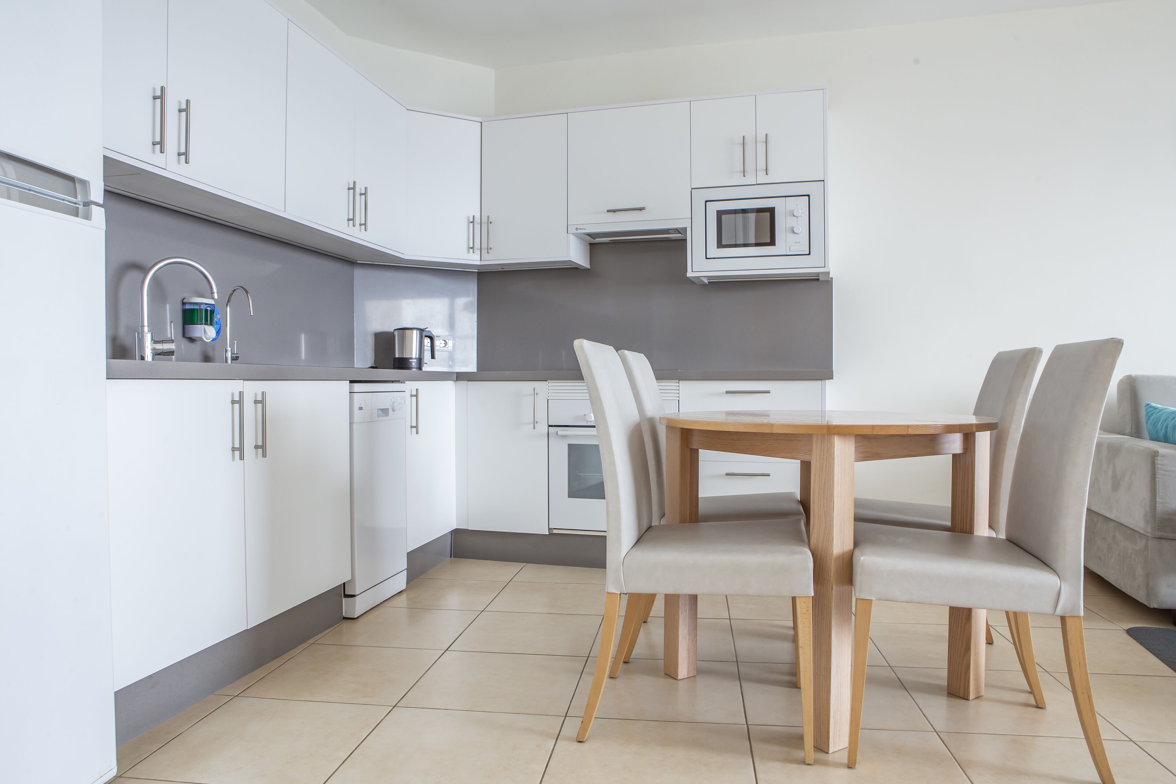A modern kitchen with white cabinets, a microwave, a kettle, and a dining table with chairs.