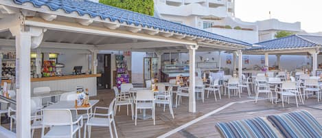 A covered outdoor seating area with white chairs and tables, a bar, and a poolside lounge with striped cushions.