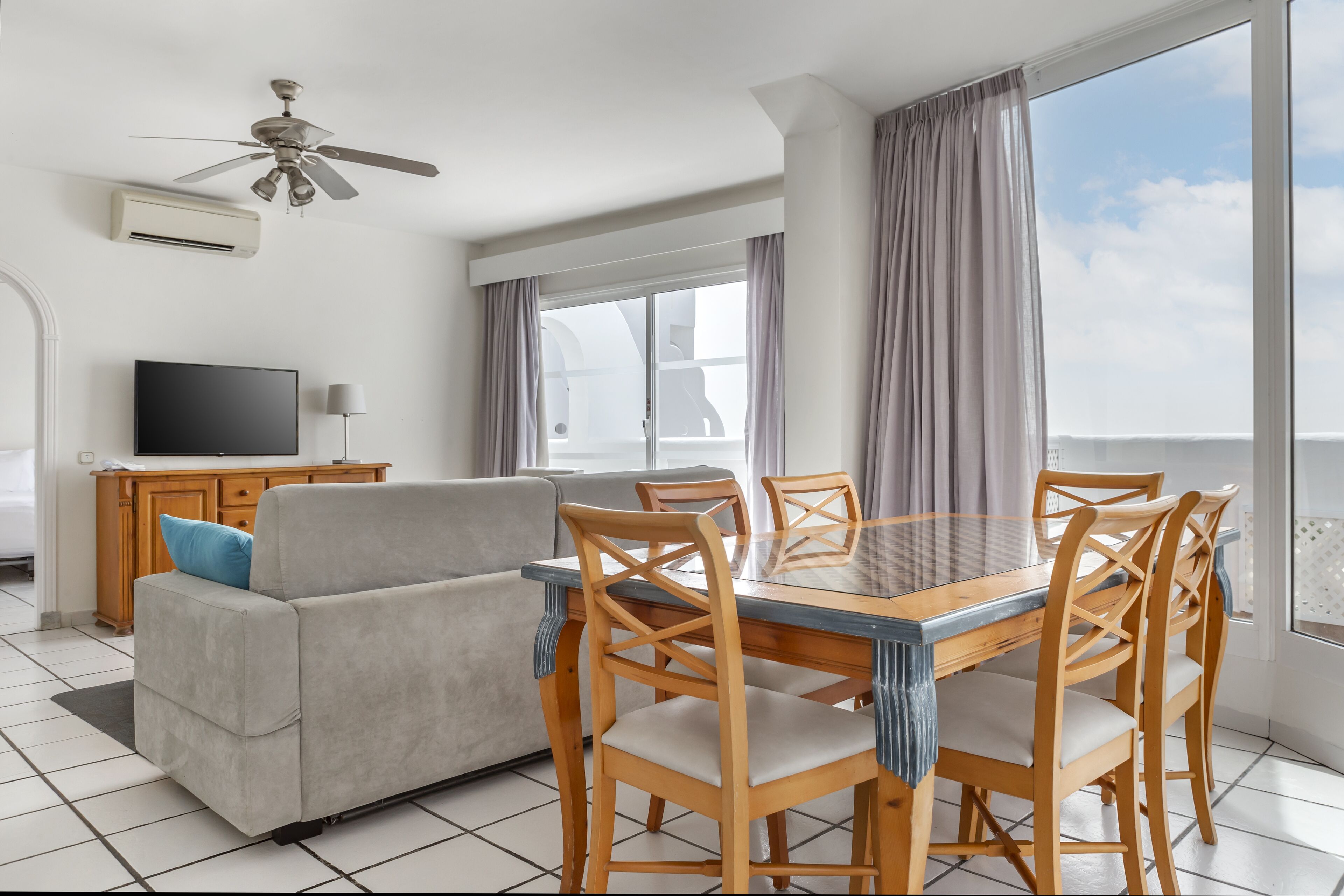 A modern dining area with a glass table and wooden chairs, a sofa, a television, and a ceiling fan.