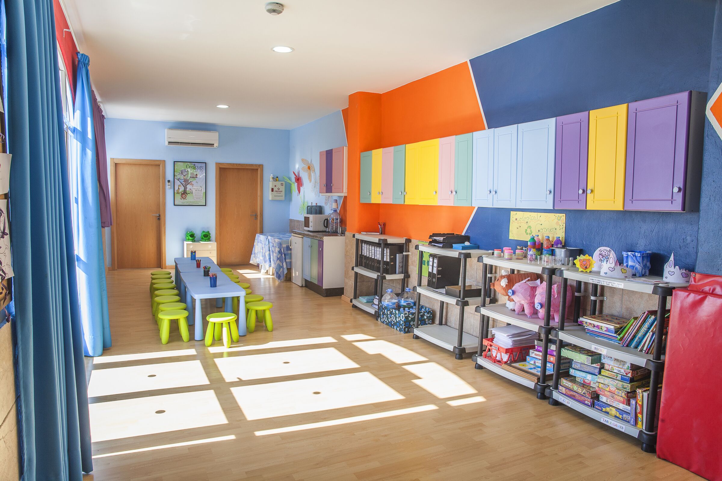 Ein helles Kinderzimmer mit einem langen Tisch, grünen Hocker und Regalen voller Spielzeug und Büchern.