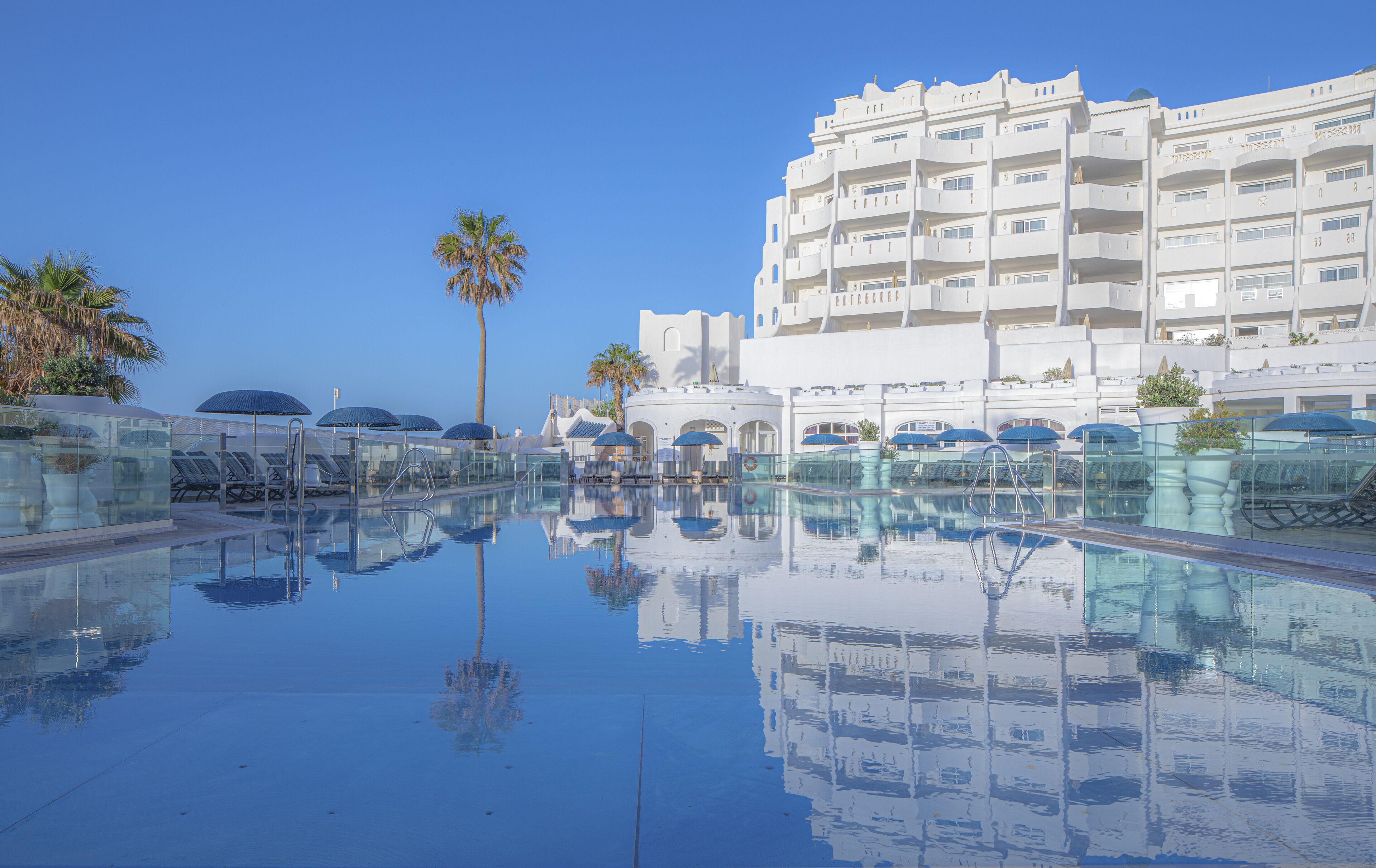 Ein Poolbereich mit klarem Wasser, das die umliegende Architektur spiegelt, darunter ein mehrstöckiges Gebäude mit Balkonen und eine Palme.