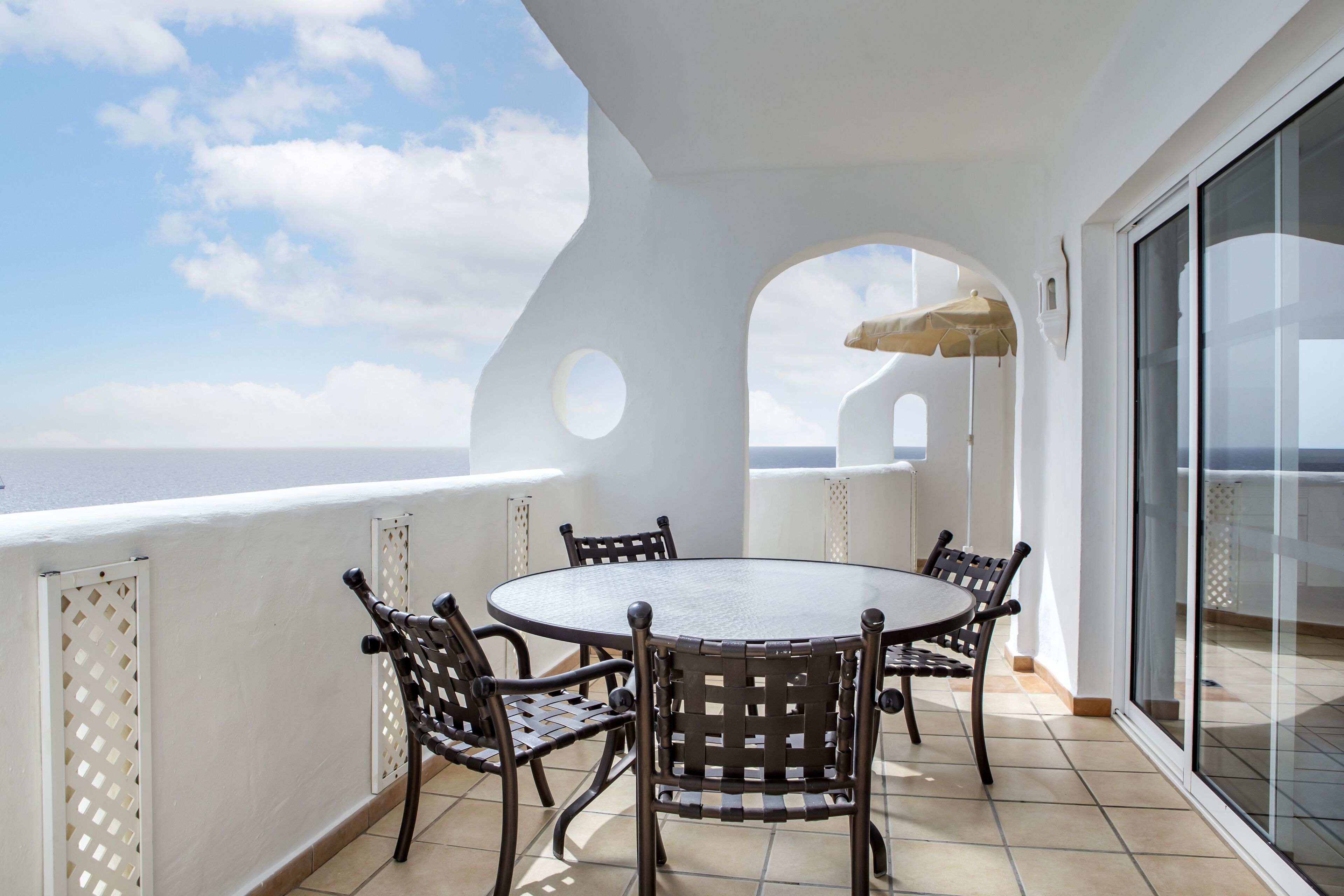 A balcony with a round table and chairs, a glass door, and a view of the sea.