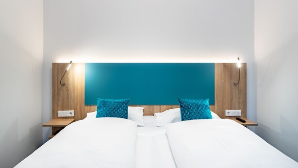 A modern hotel room with a large bed, two nightstands, a teal accent wall, and a bright overhead light. - DaunTown Rooms - Self Check-In (Vienna)