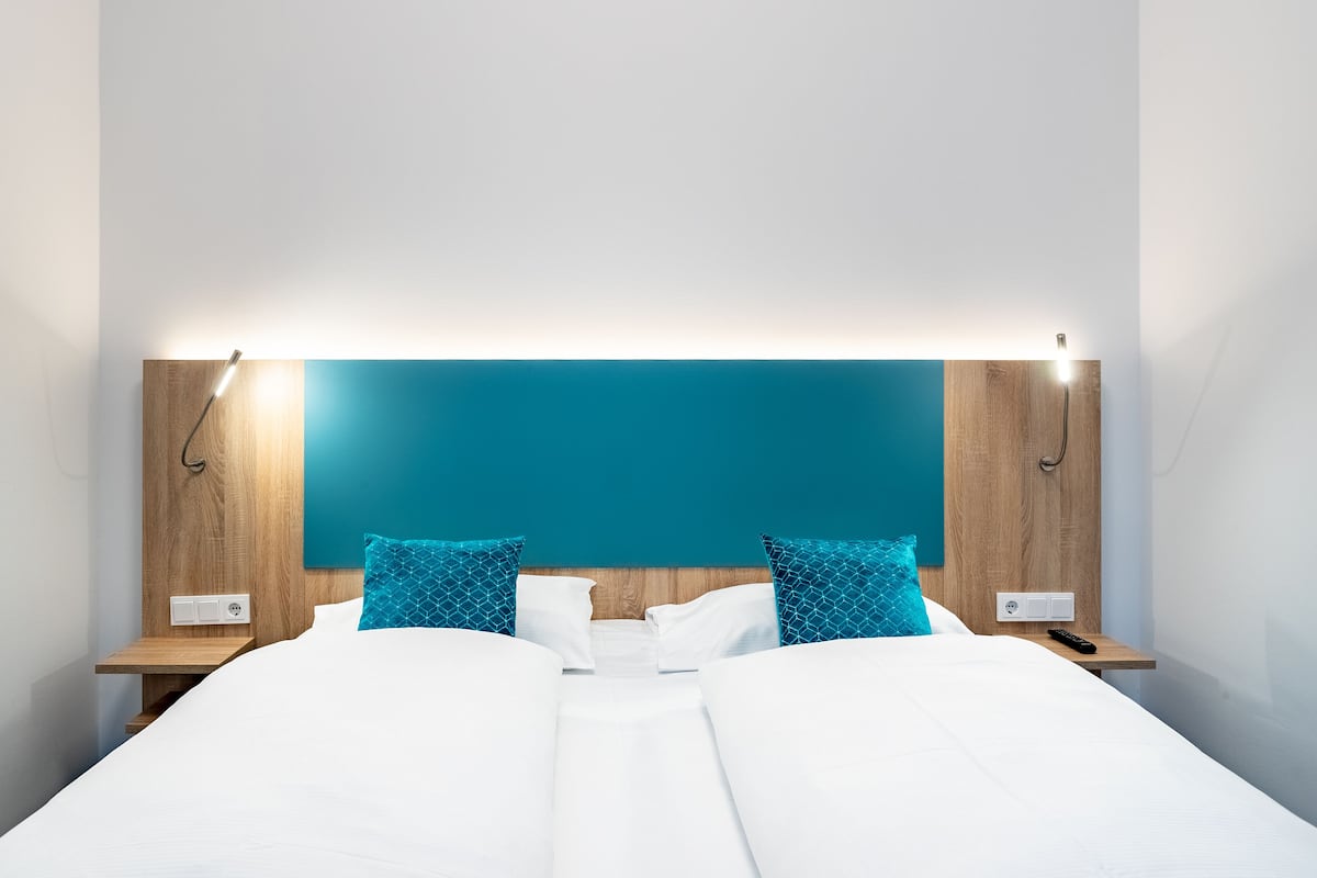 A modern hotel room with a large bed, two nightstands, a teal accent wall, and a bright overhead light.