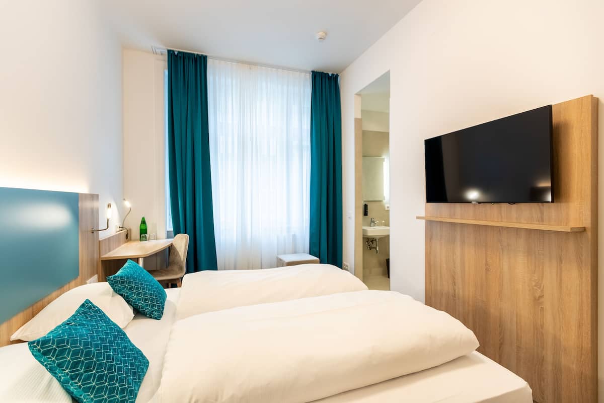 A hotel room with two beds, a TV mounted on the wall, a desk with a chair, and a bathroom visible through an open door.