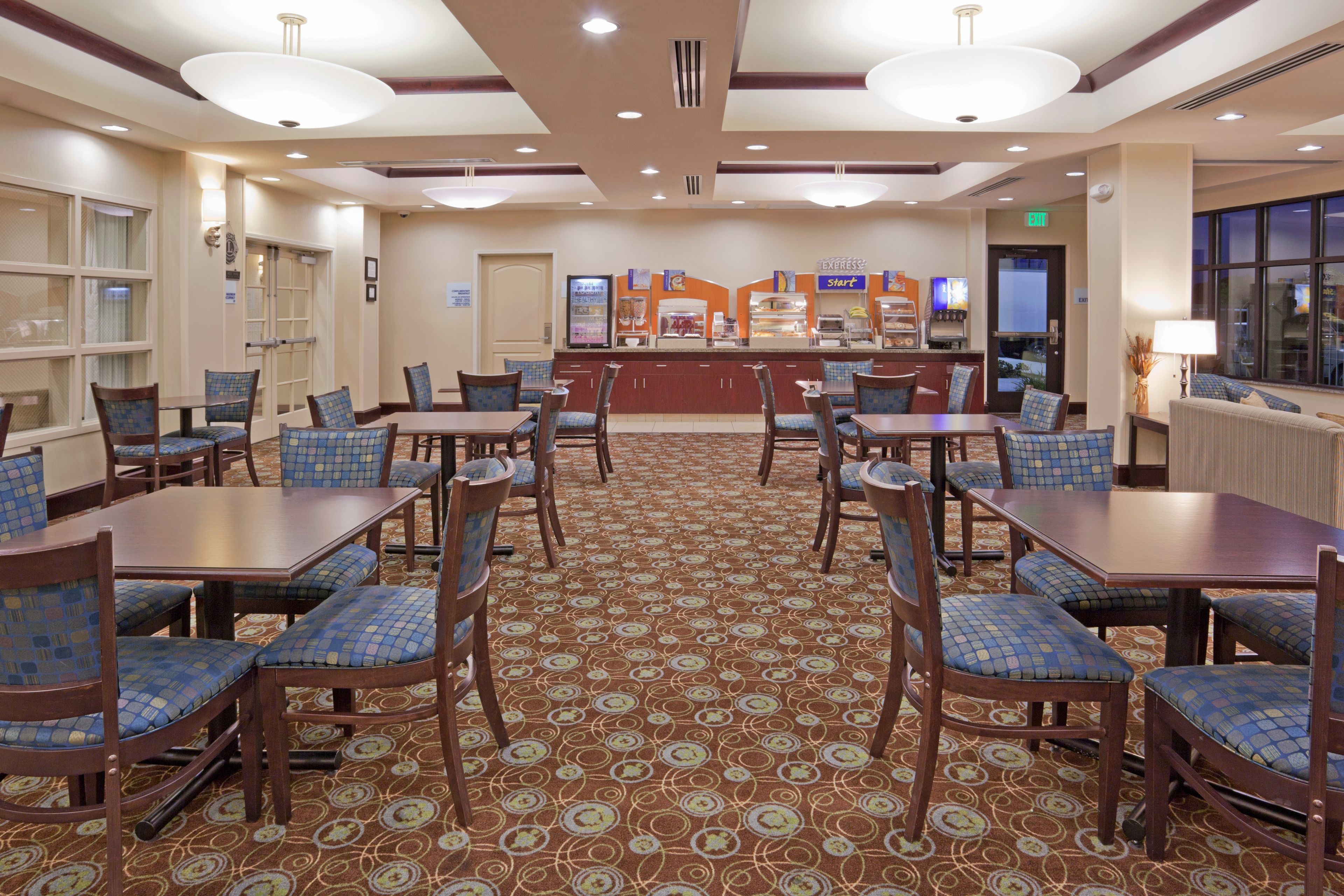 A hotel dining area with multiple tables and chairs, a central buffet, and a patterned carpet.