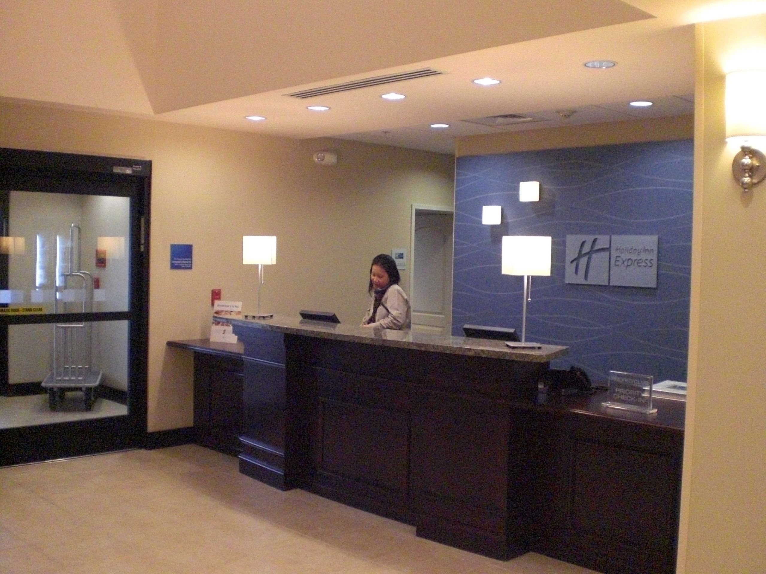 Un mostrador de recepción de hotel con una recepcionista, una pared azul con un logo y un cartel de Holiday Inn Express.