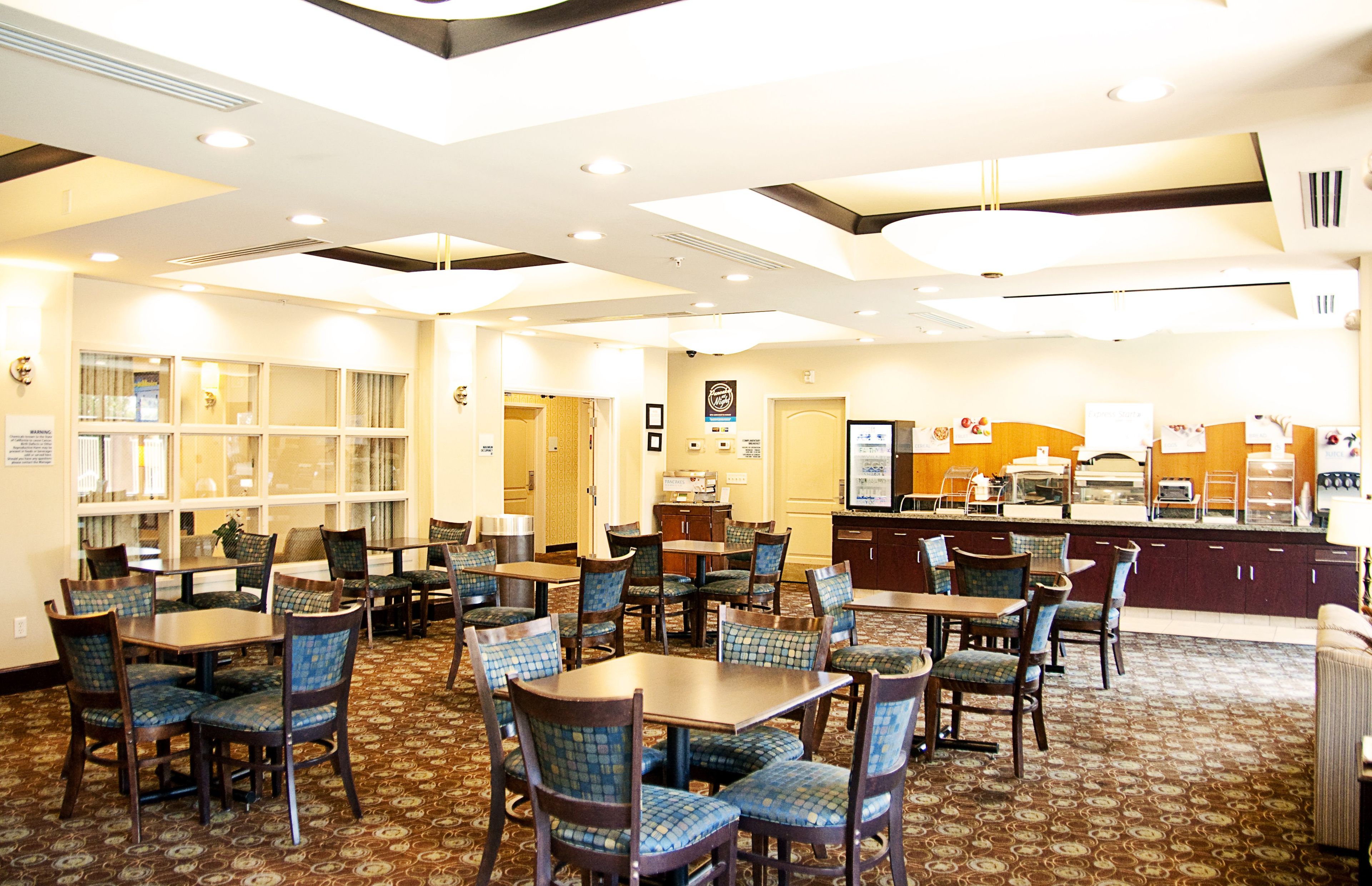 A hotel dining area with multiple tables and chairs, a counter with a menu display, and a patterned carpet.