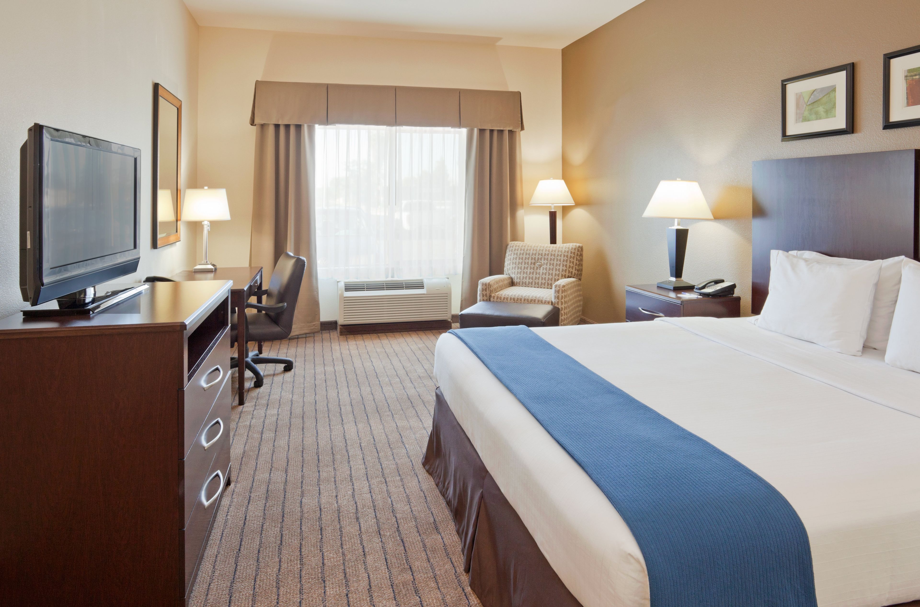 Ein Hotelzimmer mit einem Bett, einem Schreibtisch mit Fernseher, einem Sessel, einem Nachttisch mit Lampe und einer Klimaanlage.