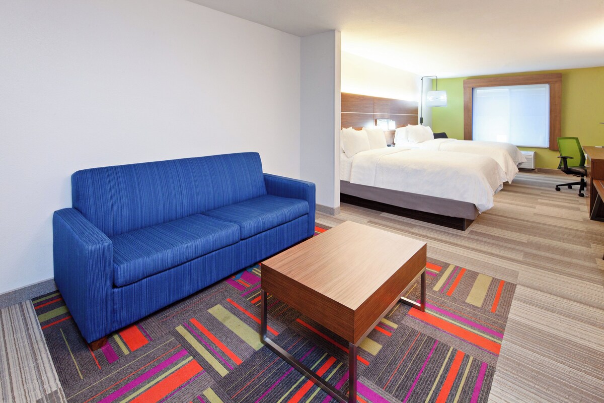 A hotel room with a blue sofa, a small wooden table, a bed with white linens, a green chair, and a colorful striped carpet.