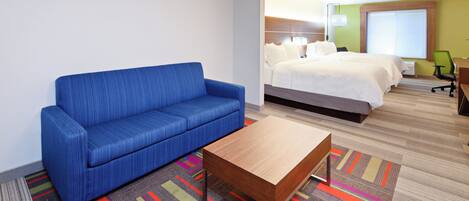 Habitación de hotel con sofá azul, cama con ropa blanca, una mesita de madera y una alfombra a rayas de colores.